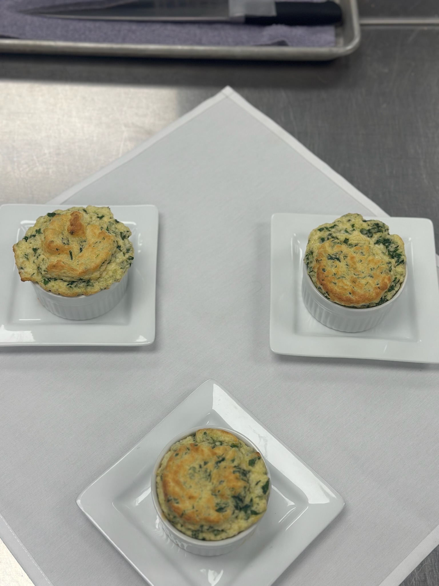 Three spinach soufflés in ramekins on white plates, displayed on a table in a kitchen setting.